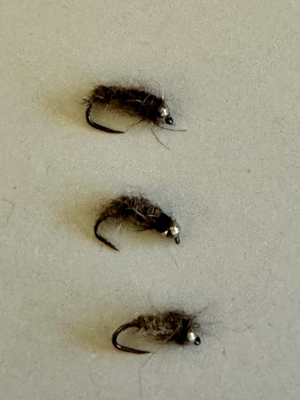 Three brown, fuzzy-bodied flies with curved hooks and metallic beads, arranged on a light surface.