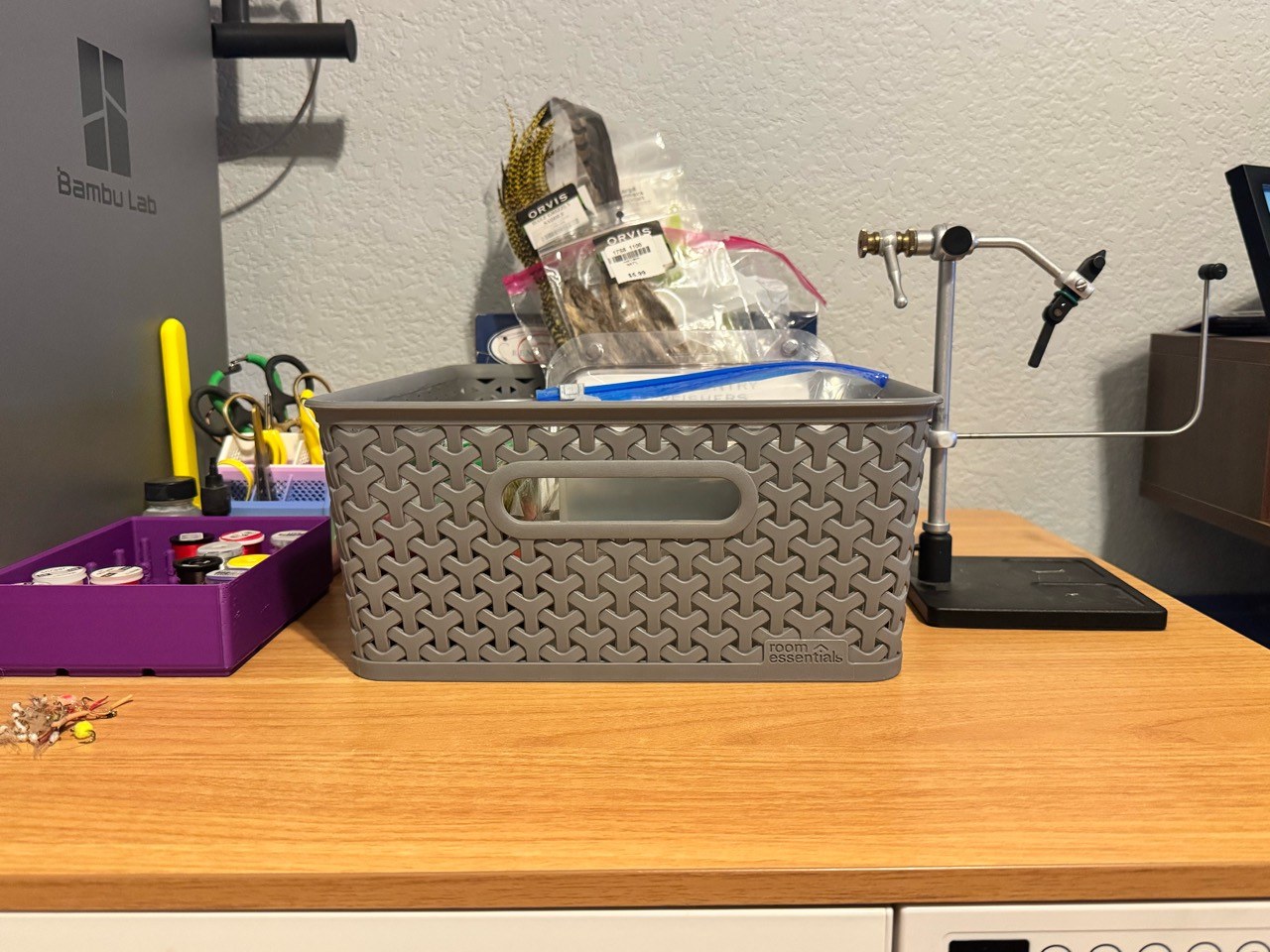 A fly-tying vise beside a gray basket of bagged materials and a purple tray of thread spools on a wooden desk.